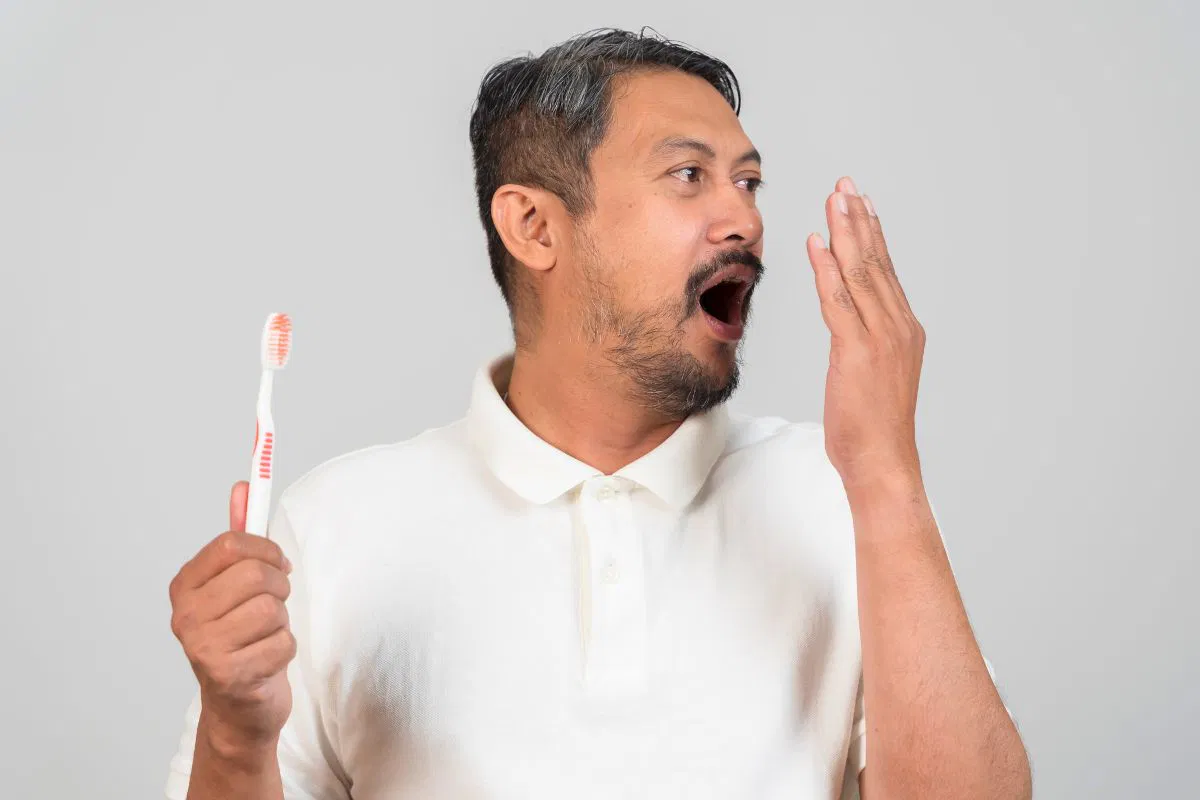 Man checking his breath with a toothbrush in hand, wondering why his breath smells after brushing.
