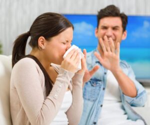 Couple sitting on a couch as the woman sneezes into a tissue while her partner covers his face, depicting shared frustration from gustatory rhinitis episodes.