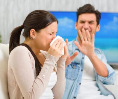 Couple sitting on a couch as the woman sneezes into a tissue while her partner covers his face, depicting shared frustration from gustatory rhinitis episodes.