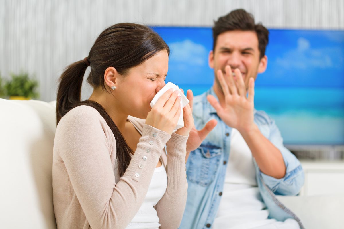 Couple sitting on a couch as the woman sneezes into a tissue while her partner covers his face, depicting shared frustration from gustatory rhinitis episodes.