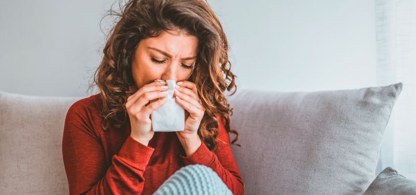 Woman in red sweater on a couch holding a tissue to her nose while sneezing, illustrating symptoms of gustatory rhinitis triggered by eating or spicy foods.