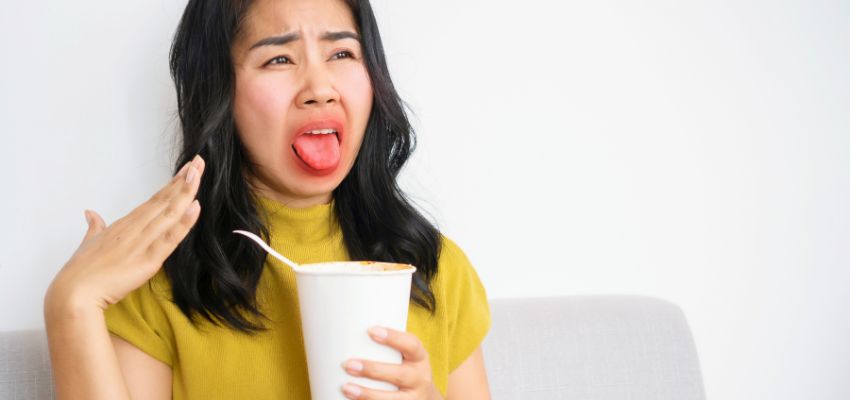 Young woman in yellow top making a face while tasting hot soup from a to-go cup, reacting to gustatory rhinitis with sudden nasal irritation and runny nose.
