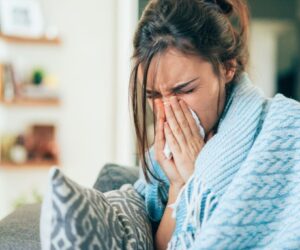 Woman wrapped in a blanket sitting on a couch using tissue to blow her nose while dealing with flu symptoms, illustrating the need to know how to cure flu fast at home.