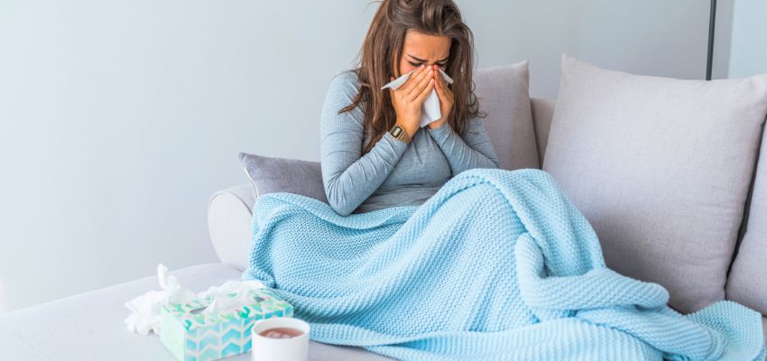 Woman curled up on a couch under a blue blanket, holding tissue to her face during a severe sneeze, highlighting discomfort and the desire to learn how to cure flu fast.