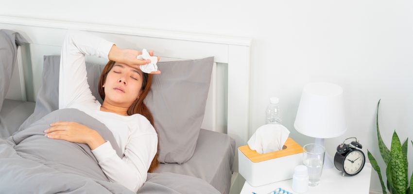 Woman lying in bed with eyes closed, touching her forehead with a tissue while resting under a blanket, showing common flu recovery steps and how to cure flu fast with rest.