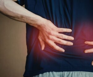 Side view of a man in a dark blue t-shirt standing and placing both hands on his lower and mid-back with a red glow highlighting the area of pain, conveying referred or radiating discomfort that prompts many to ask 'can gerd cause back pain' during episodes of severe reflux