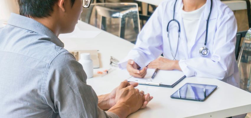 Male patient in a gray shirt seated at a desk across from a female physician in a white coat with stethoscope, both engaged in serious conversation with clipboard and tablet nearby, representing a medical consultation where patients often inquire 'can gerd cause back pain' as part of evaluating gastrointestinal symptoms and referred pain patterns