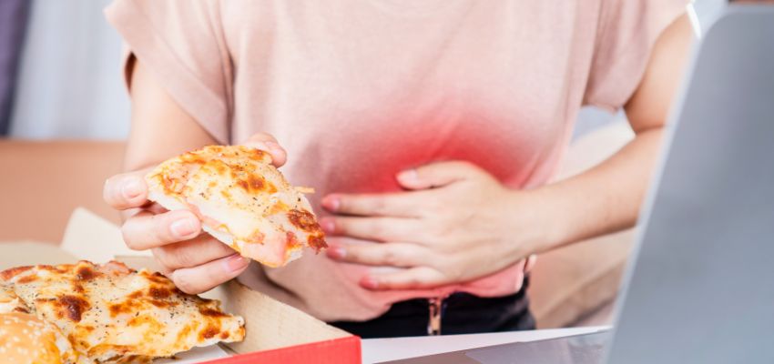 Person in casual clothing experiencing discomfort, holding a slice of pizza in one hand and pressing the other hand against their upper stomach highlighted in red, clearly depicting the pain and triggers commonly associated with a GERD flare up after eating